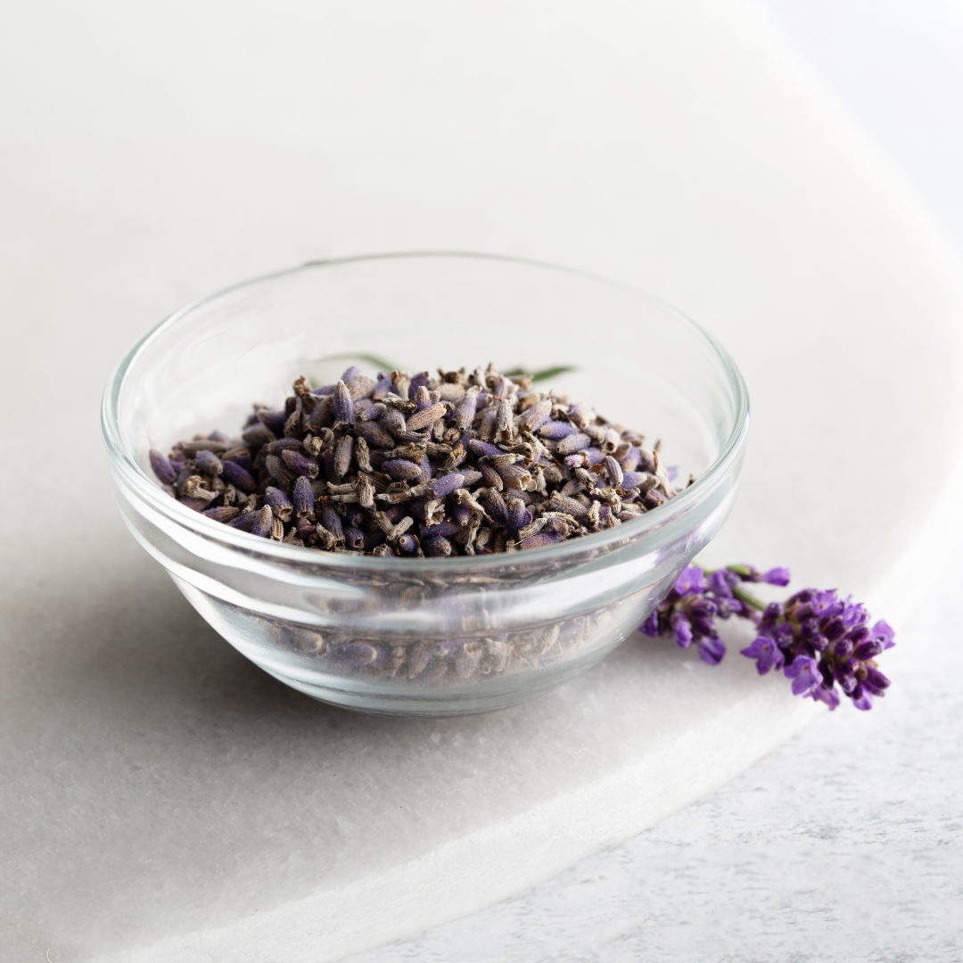 Dried lavender heads in a clear glass bowl with a fresh sprig of lavender which accompanies article on the skin benefits of lavender
